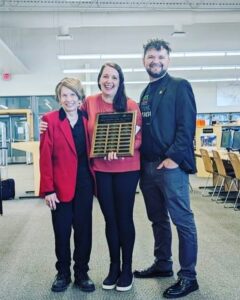 Image of three people standing together and smiling in a classroom setting. The person in the center is holding the KLL Arizona Teacher of the Year award.